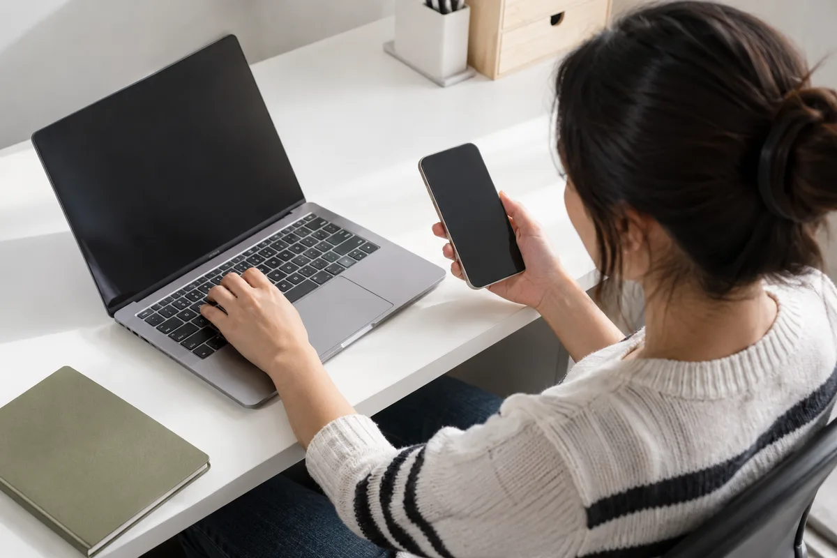 Manos utilizando computadora y teléfono con pantallas fuera de vista.